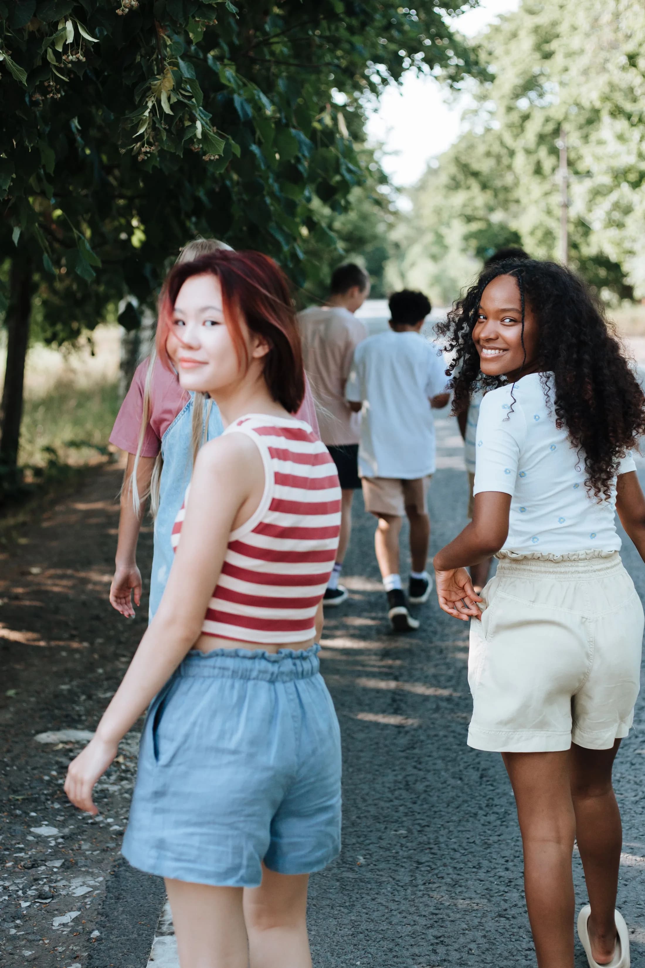A group of young people walking together down a tree-lined lane, two of them glancing back at the camera.